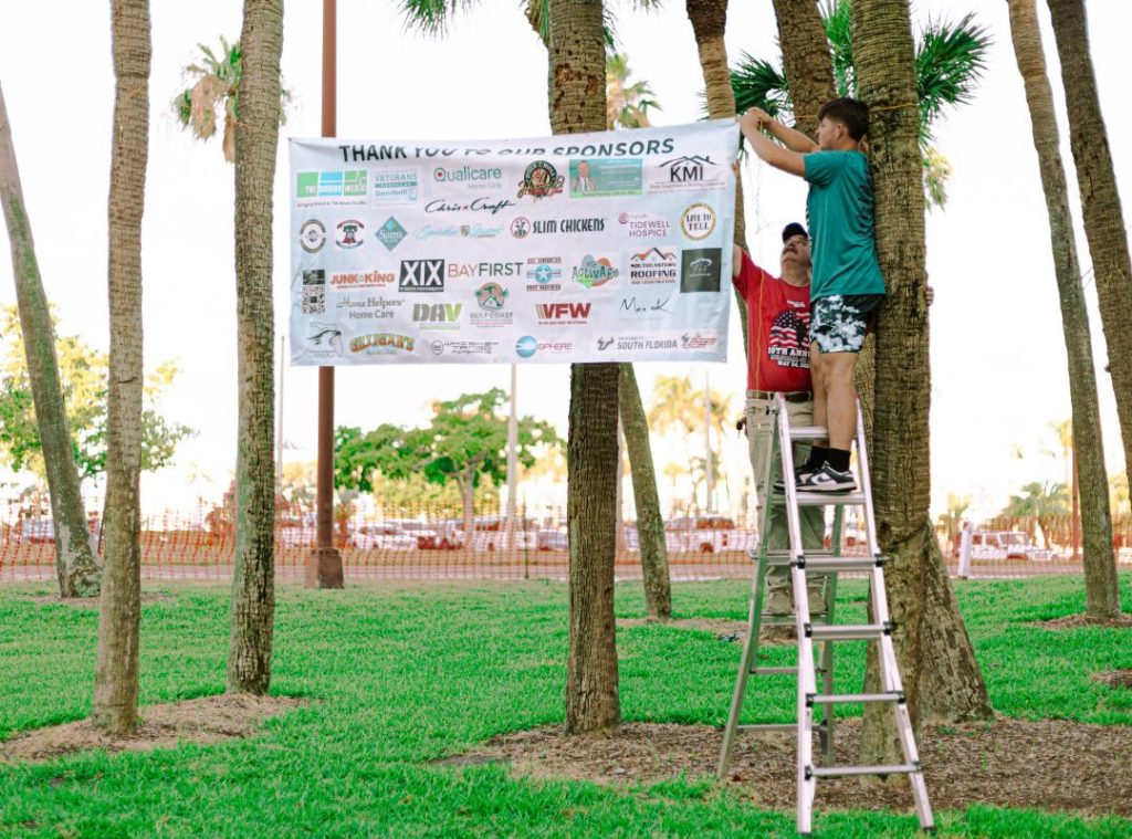 Volunteers hanging a Thank You to Our Sponsors banner between palm trees at a Sarasota park before the SRQ Vets Annual Memorial Hike, showcasing local business support for the veteran community.