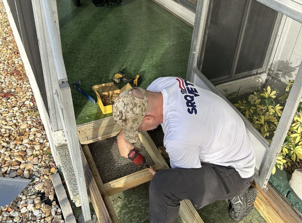An SRQ Vets volunteer in a branded white shirt and military cap using a power drill to install a mobility ramp at a Sarasota home, demonstrating the hands-on veteran community support SRQ Vets provides.