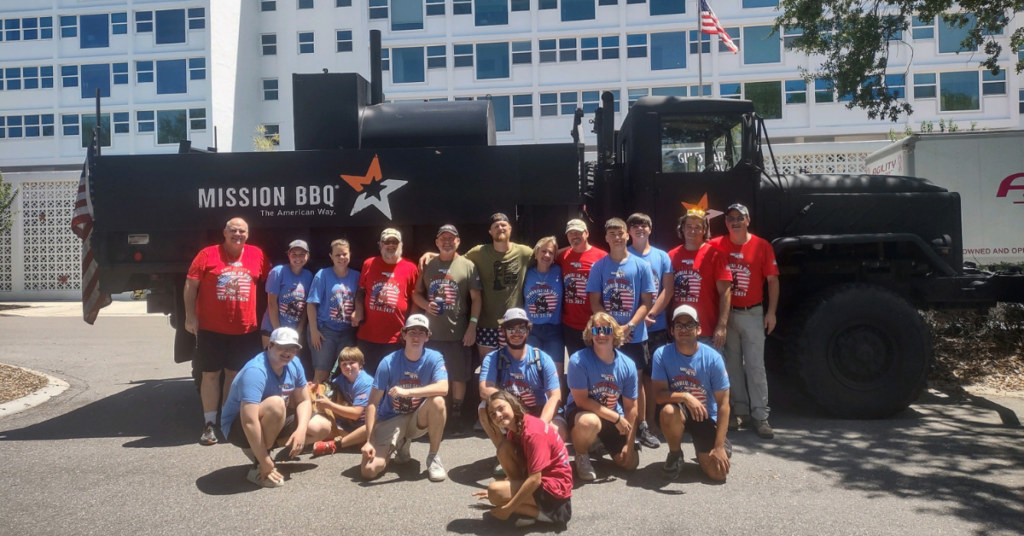 A large group of SRQ Vets participants and volunteers posing together in front of a Mission BBQ truck at the Annual Memorial Hike in Sarasota, Florida, showing the community support network behind Lakewood Ranch and Suncoast veterans.